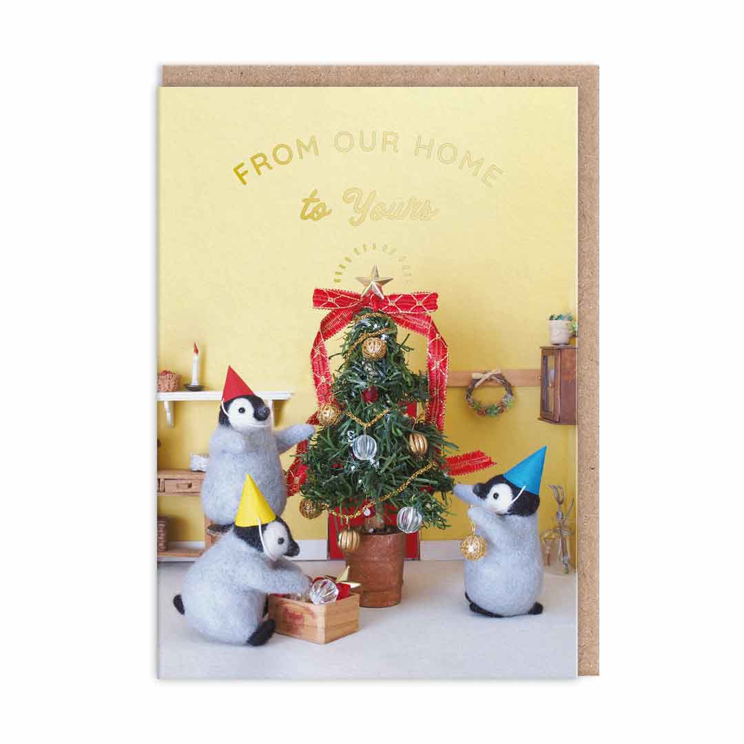 TChristmas card featuring an image of toy penguins dressing a Christmas tree. Gold foil text reads "From Our Home To Yours"